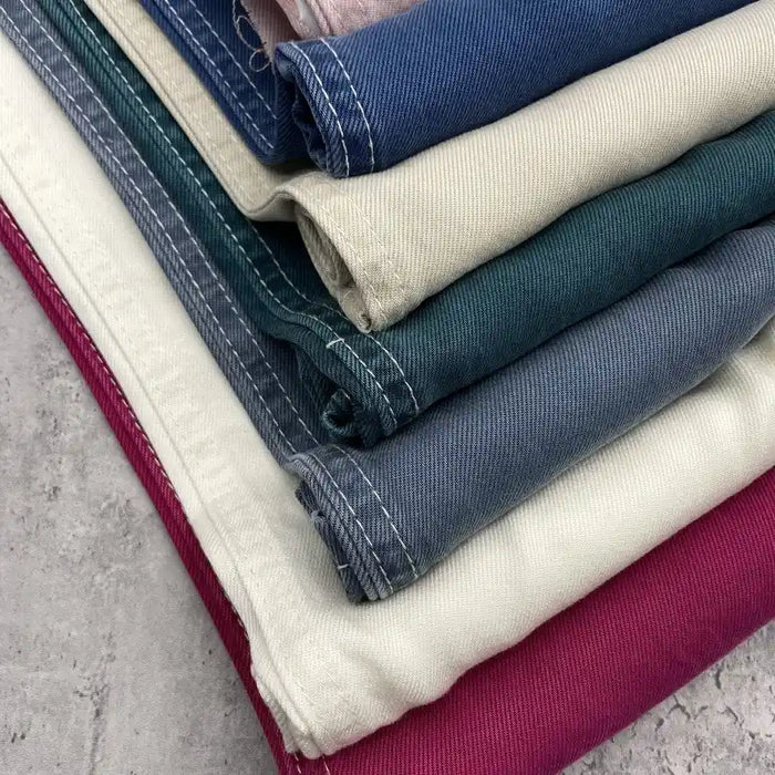 A stack of Solid Color Denim Lyocell Fabric by the yard from Longan Craft in cream, dark green, blue, and magenta with white edge stitching—wrinkle-free, breathable, and perfect for sustainable jeans and denim apparel.