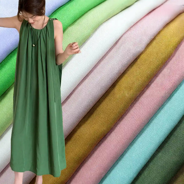 A woman in a loose green dress stands before neatly stacked rolls of Longan Craft Solid Color Linen Lyocell Fabric By The Yard—sustainable, wrinkle-free textiles in green, white, pink, gold, and blue for dresses.