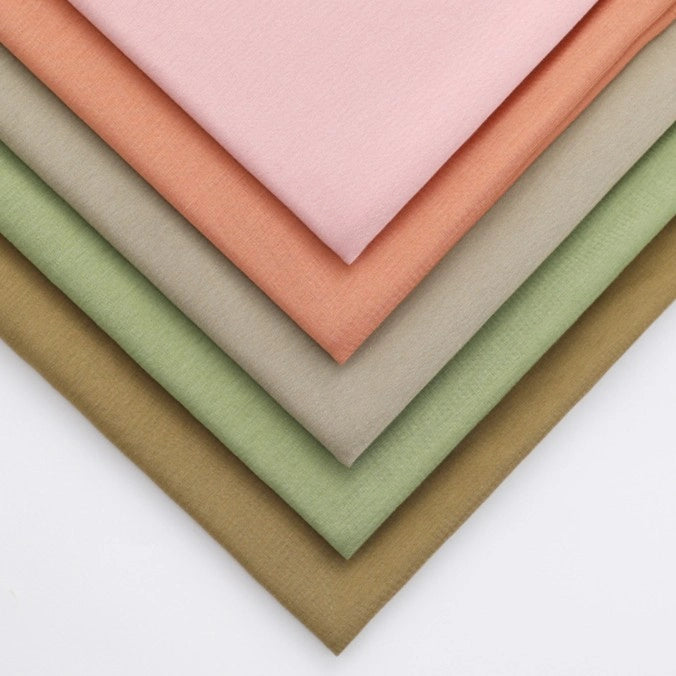 Five overlapping pieces of Longan Craft Solid Color Modal Jersey Cotton Spandex Fabric in pastel pink, peach, beige, light green, and olive are neatly fanned on a white surface—ideal for sleepwear and children’s clothing projects.