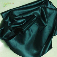 A piece of Longan Craft Solid Color Polyester Satin Fabric, in dark green with a silky stretch satin texture, lies slightly wrinkled on a light surface, showcasing its smooth and lustrous finish—ideal for blouses.