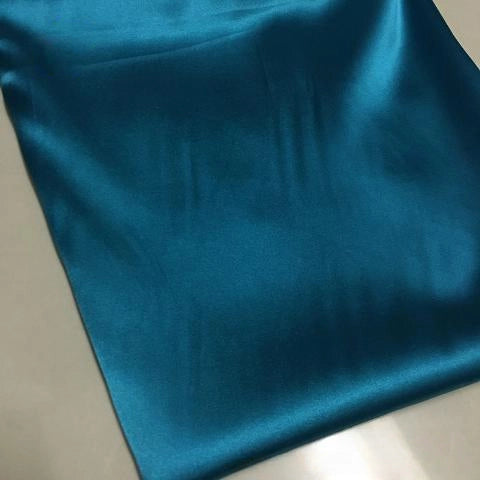 Longan Craft's Solid Color Polyester Satin Fabric By The Yard in rich teal has a silky stretch satin texture, a glossy finish, and is perfect for blouses. The smooth, shiny material reflects light on a flat, light-colored surface.