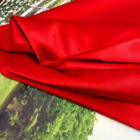 A piece of Longan Craft Solid Color Polyester Satin Fabric By The Yard in red, known for its silky stretch satin texture, is draped over a surface with greenery and a white fence, catching the light and showing off its smooth, glossy finish.