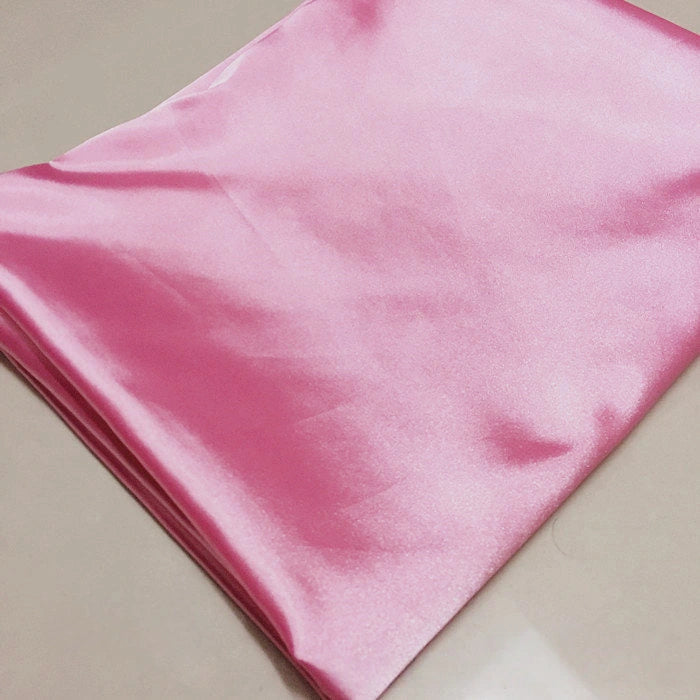A neatly folded piece of Longan Craft Solid Color Polyester Satin Fabric in pink, known for its silky stretch texture and perfect for blouses, is displayed on a smooth beige surface.