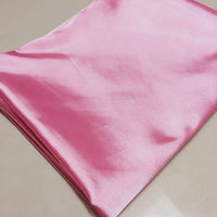 A neatly folded piece of Longan Craft Solid Color Polyester Satin Fabric in pink, known for its silky stretch texture and perfect for blouses, is displayed on a smooth beige surface.