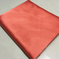 A piece of Longan Craft Solid Color Polyester Satin Fabric in coral, featuring a silky stretch satin texture, rests on a light surface, highlighting its smooth and lustrous appeal—perfect material for blouses.