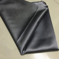 A piece of Longan Craft Solid Color Polyester Satin Fabric By The Yard, featuring a silky stretch satin texture in solid black, is spread on a light surface, reflecting light and highlighting its soft, glossy sheen—ideal for blouses.