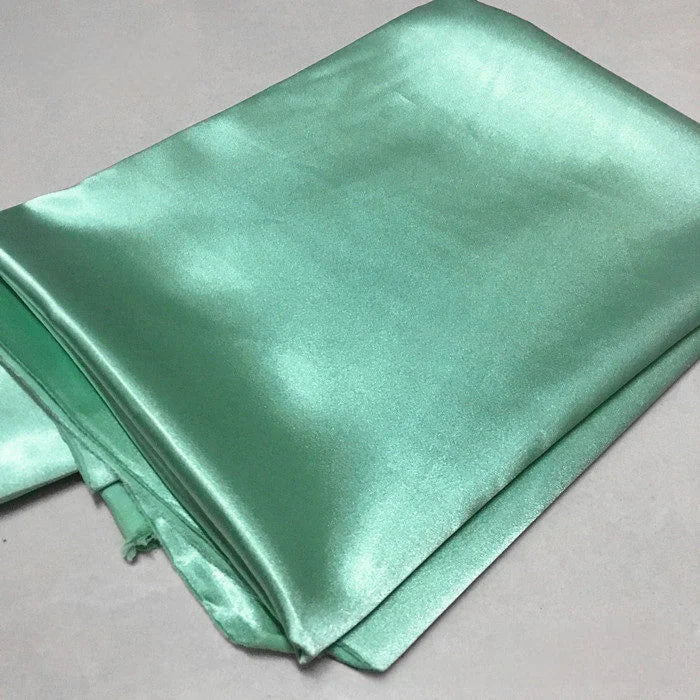 A piece of Longan Craft Solid Color Polyester Satin Fabric By The Yard in shiny light green with a silky stretch texture is folded on a light gray surface, ideal for blouses.