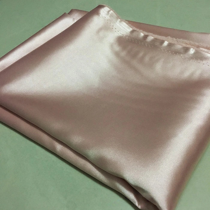 A piece of Longan Craft Solid Color Polyester Satin Fabric By The Yard, featuring a silky stretch satin texture in pale pink, with a smooth, lustrous surface, is displayed on a light-colored background—ideal for blouses.