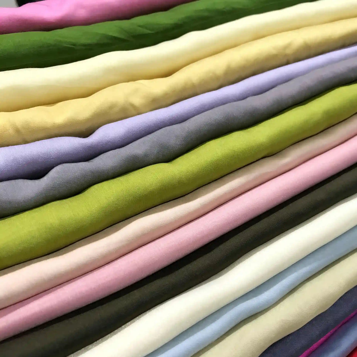 Neatly stacked rolls of Longan Craft Solid Color Ramie Fabric by the yard, available in an array of hues including green, cream, yellow, purple, pink, blue, and brown are arranged in horizontal layers. This super lightweight lyocell ramie material is breathable and perfect for blouses, offering both vibrant color and comfort.