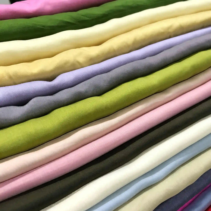 Neatly stacked rolls of Longan Craft Solid Color Ramie Fabric by the yard, available in an array of hues including green, cream, yellow, purple, pink, blue, and brown are arranged in horizontal layers. This super lightweight lyocell ramie material is breathable and perfect for blouses, offering both vibrant color and comfort.