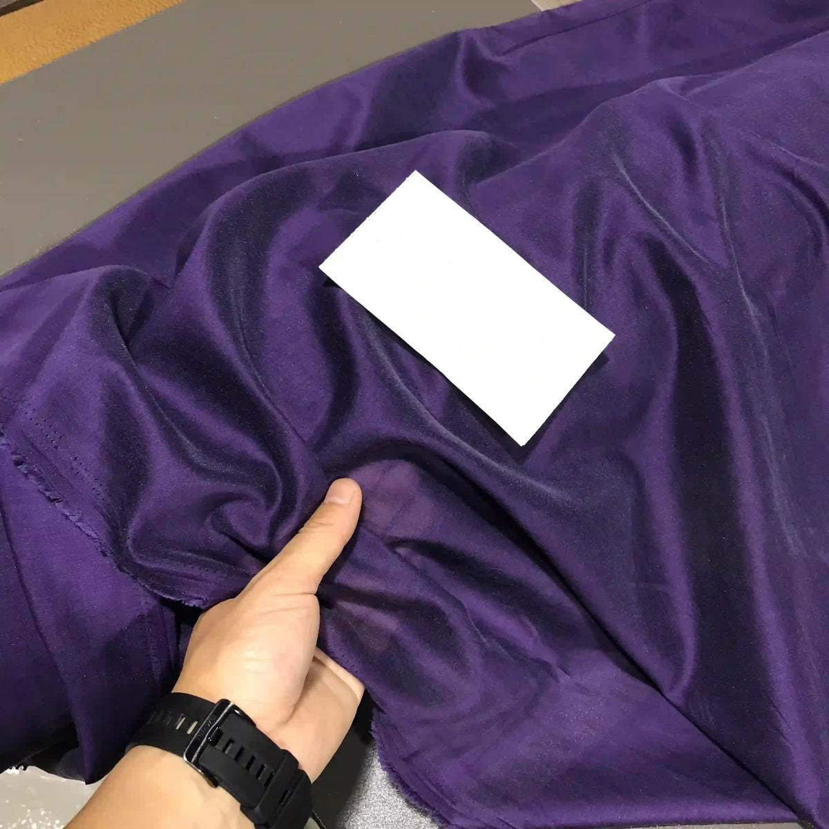 A hand wearing a black watch holds a piece of shiny purple Solid Color Ramie Fabric by the Yard from Longan Craft, a super lightweight lyocell ramie material known for its breathable texture, ideal for blouses. A blank white card rests on top of the fabric.