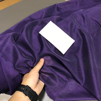 A hand wearing a black watch holds a piece of shiny purple Solid Color Ramie Fabric by the Yard from Longan Craft, a super lightweight lyocell ramie material known for its breathable texture, ideal for blouses. A blank white card rests on top of the fabric.
