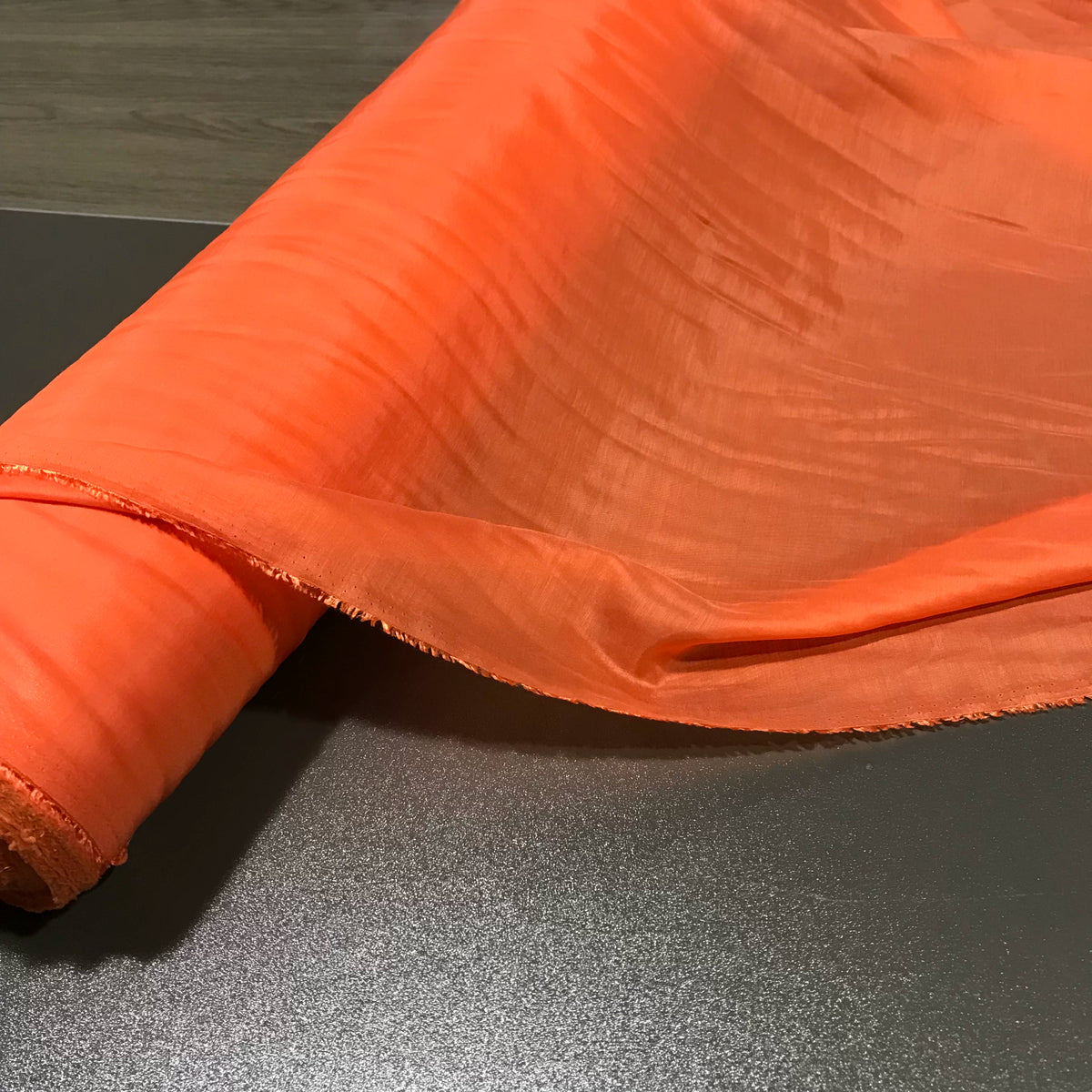 A roll of Longan Craft’s Solid Color Ramie Fabric By The Yard, a super lightweight lyocell ramie material in bright orange, is partially unrolled on a dark gray surface. This breathable fabric for blouses features a subtle sheen and visible texture lines.