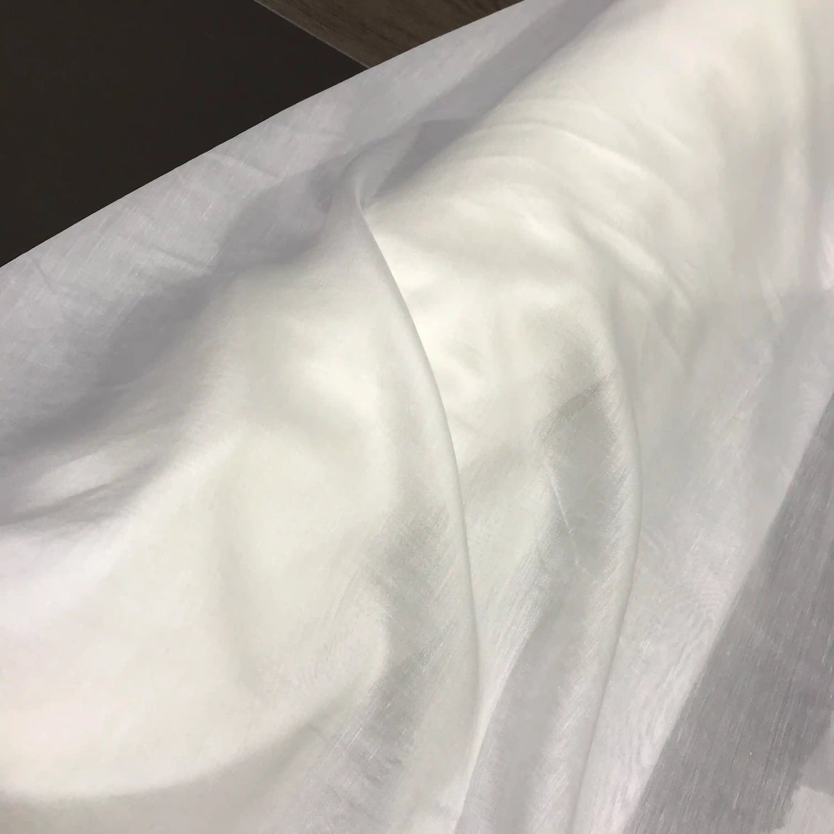 A length of Longan Craft’s Solid Color Ramie Fabric by the Yard, super lightweight and semi-sheer with a lyocell ramie blend, drapes smoothly over a surface, forming gentle folds and soft shadows. This breathable material adds an airy lightness and refined elegance—ideal for blouses and delicate projects.