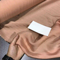 A hand holds a piece of light peach-colored Solid Color Ramie Fabric By The Yard from Longan Craft, a super lightweight lyocell ramie material for blouses, displayed on a table with a white rectangular card placed on top.