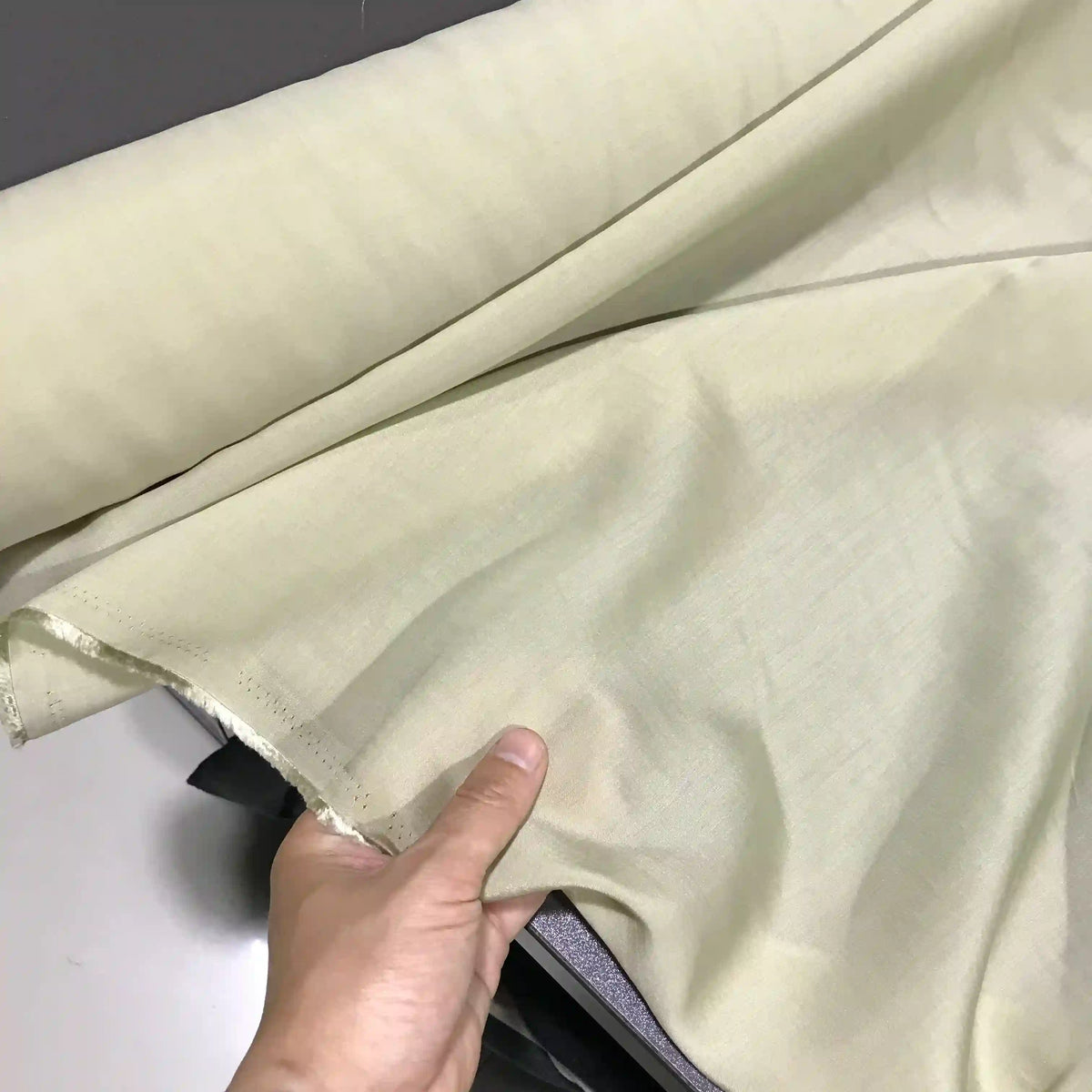 A hand holds up a piece of Solid Color Ramie Fabric By The Yard from Longan Craft, showcasing its super lightweight, breathable lyocell ramie material in light beige and highlighting its smooth texture and slightly sheer appearance—ideal for blouses.