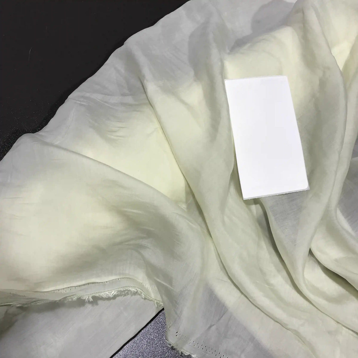 A piece of sheer, pale Solid Color Ramie Fabric by the Yard from Longan Craft, known for its super lightweight and breathable lyocell ramie material ideal for blouses, lies crumpled on a dark surface with a rectangular white card placed on top.