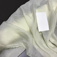 A piece of sheer, pale Solid Color Ramie Fabric by the Yard from Longan Craft, known for its super lightweight and breathable lyocell ramie material ideal for blouses, lies crumpled on a dark surface with a rectangular white card placed on top.