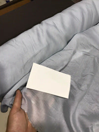 A hand holds the Solid Color Ramie Fabric By The Yard from Longan Craft in light blue, comparing its shade to a white rectangular card placed on top. The super lightweight lyocell ramie material appears soft, slightly wrinkled, and clearly showcases its breathable quality—ideal for blouses.