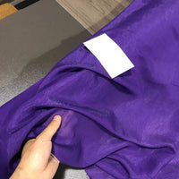 A hand holds a piece of sheer, vibrant purple Longan Craft Solid Color Ramie Fabric By The Yard—super lightweight lyocell ramie material for blouses—on a table, with a white rectangular label resting on the fabric.