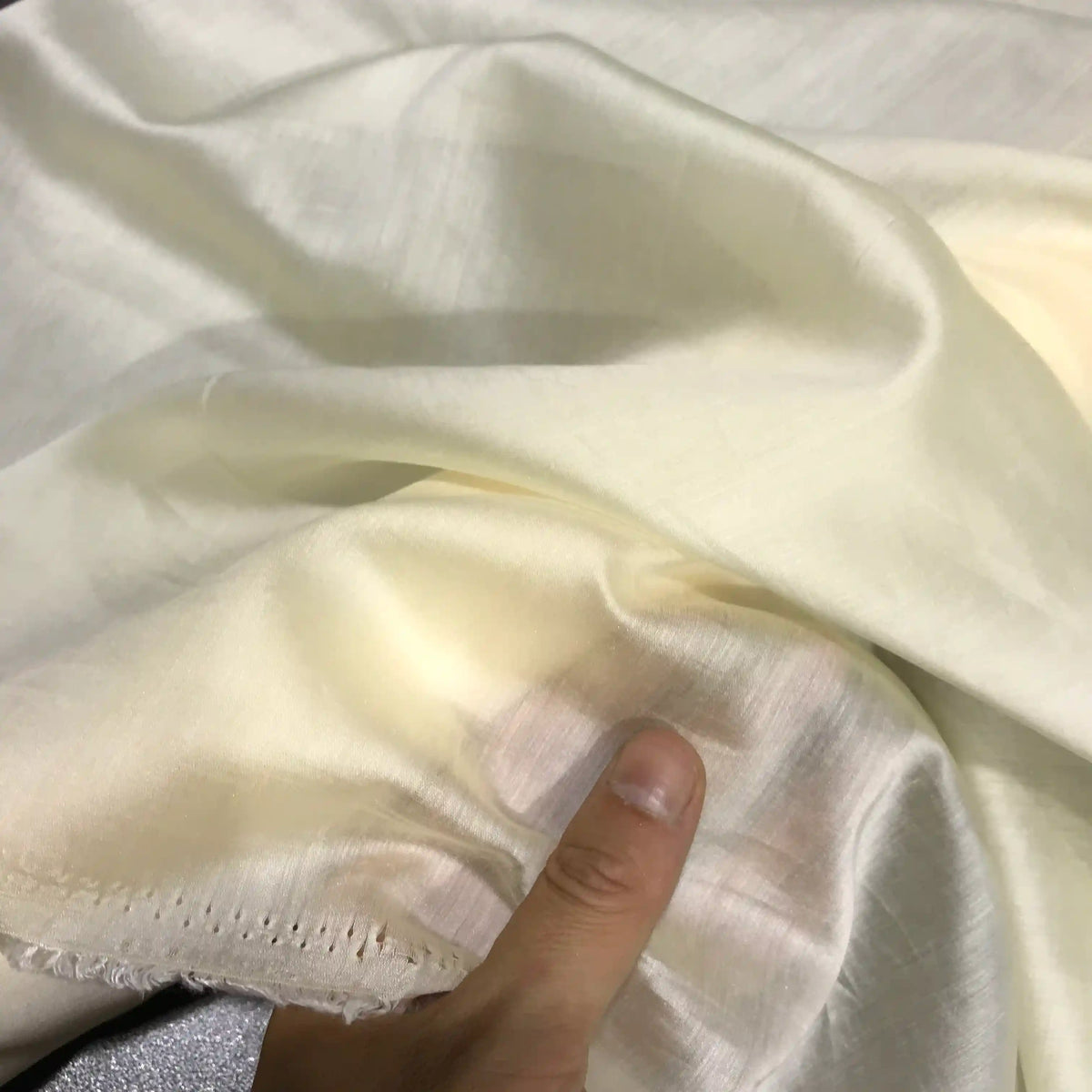 A hand holds a piece of Longan Craft Solid Color Ramie Fabric By The Yard, a super lightweight lyocell ramie material with a soft, off-white hue and subtle sheen. The breathable fabric looks thin and translucent, featuring visible stitching along one edge—perfect for crafting airy blouses.
