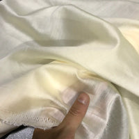 A hand holds a piece of Longan Craft Solid Color Ramie Fabric By The Yard, a super lightweight lyocell ramie material with a soft, off-white hue and subtle sheen. The breathable fabric looks thin and translucent, featuring visible stitching along one edge—perfect for crafting airy blouses.