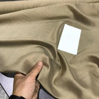 A hand is holding a piece of shiny, light brown Longan Craft Solid Color Ramie Fabric by the yard, a super lightweight, breathable lyocell ramie material perfect for blouses. A small white rectangular card rests on top of the fabric.