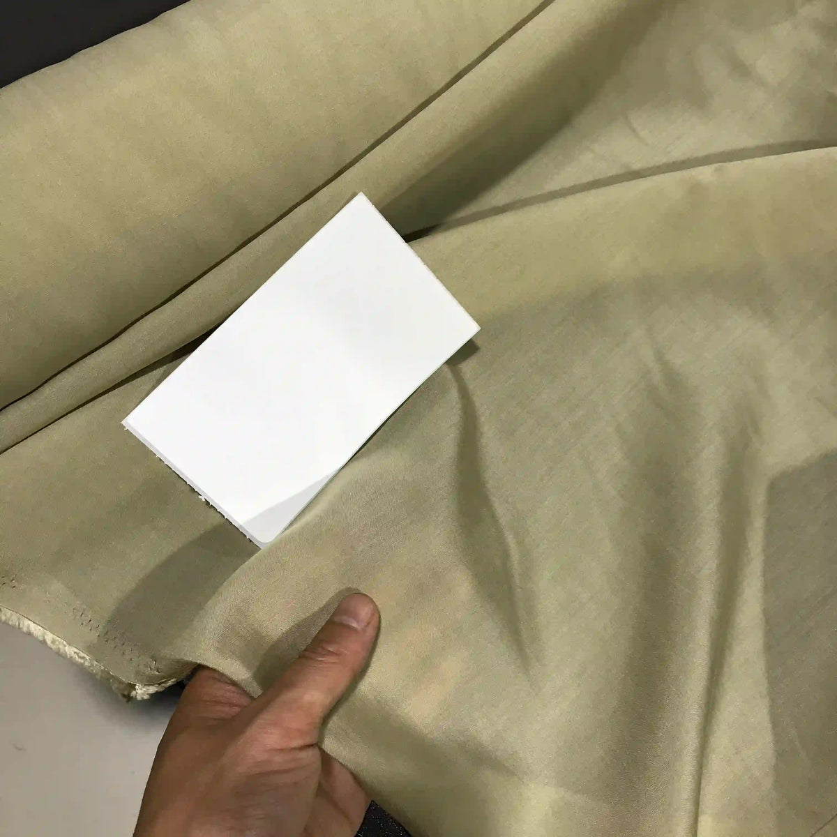 A hand displays a piece of Longan Craft’s Solid Color Ramie Fabric By The Yard, a super lightweight lyocell ramie material in light beige, with a white rectangular card placed on top for color comparison.