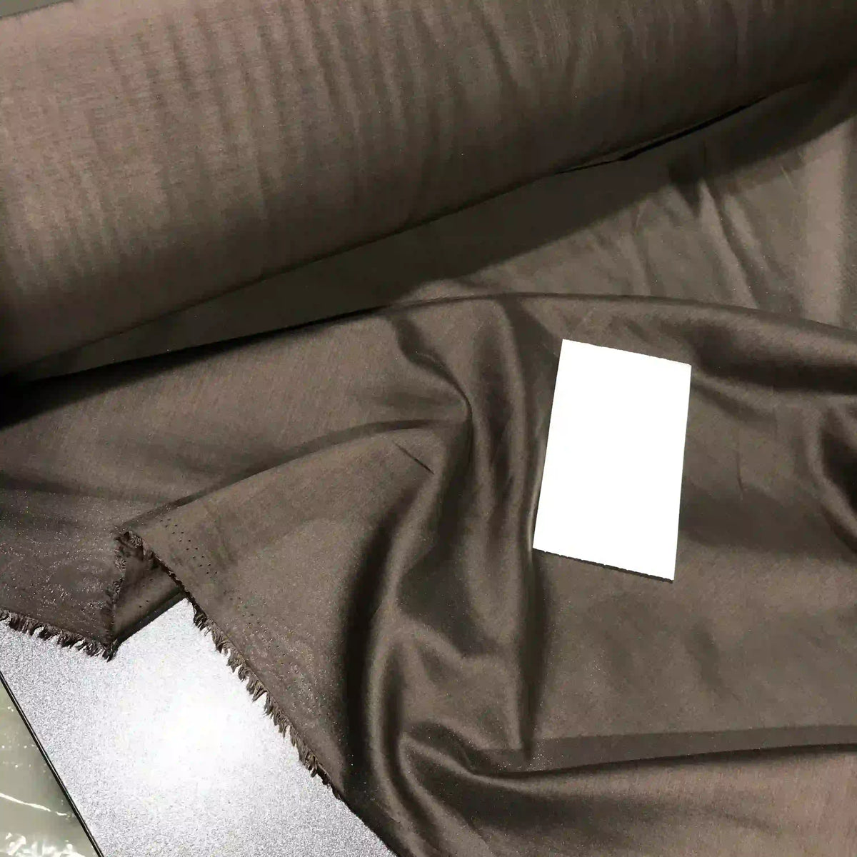 A roll of Longan Craft’s Solid Color Ramie Fabric By The Yard, shown here in a super lightweight dark brown lyocell ramie material, is partially unrolled on a flat surface with a small white rectangular card placed on the breathable fabric for scale.