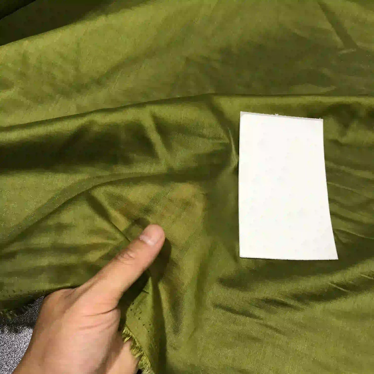 A hand displays the Solid Color Ramie Fabric By The Yard from Longan Craft—a super lightweight, breathable lyocell ramie material in green—next to a rectangular white card for texture and color comparison, ideal for blouses.