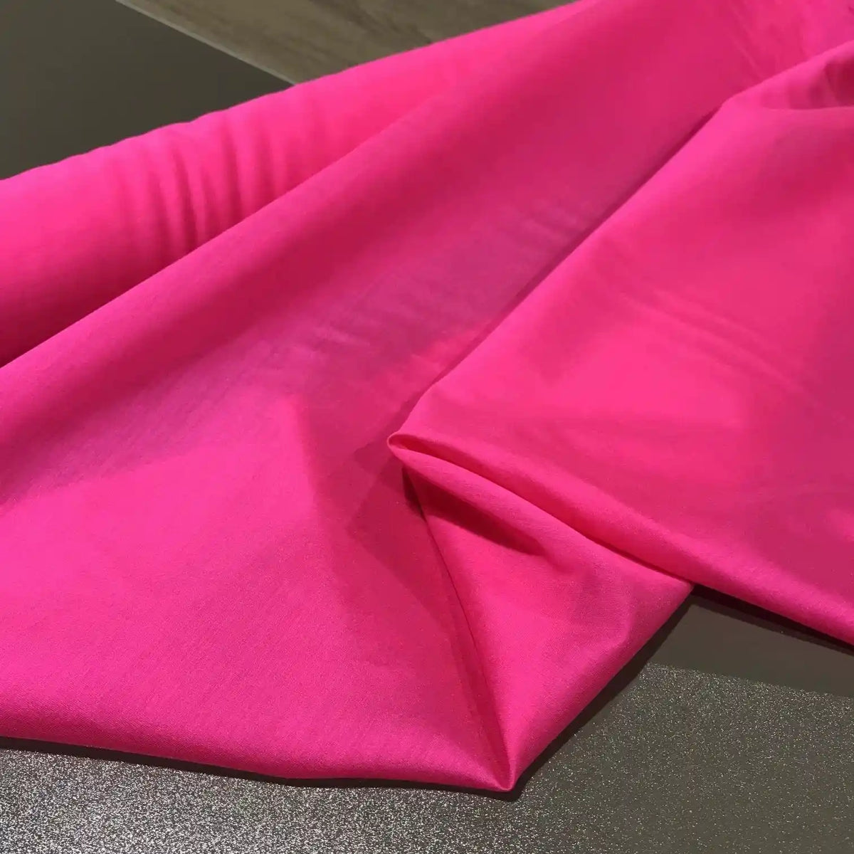 A close-up of Longan Craft’s Solid Color Ramie Fabric By The Yard, a bright pink super lightweight lyocell ramie material, draped over a dark surface and highlighting its smooth texture and subtle folds—ideal for breathable blouses.