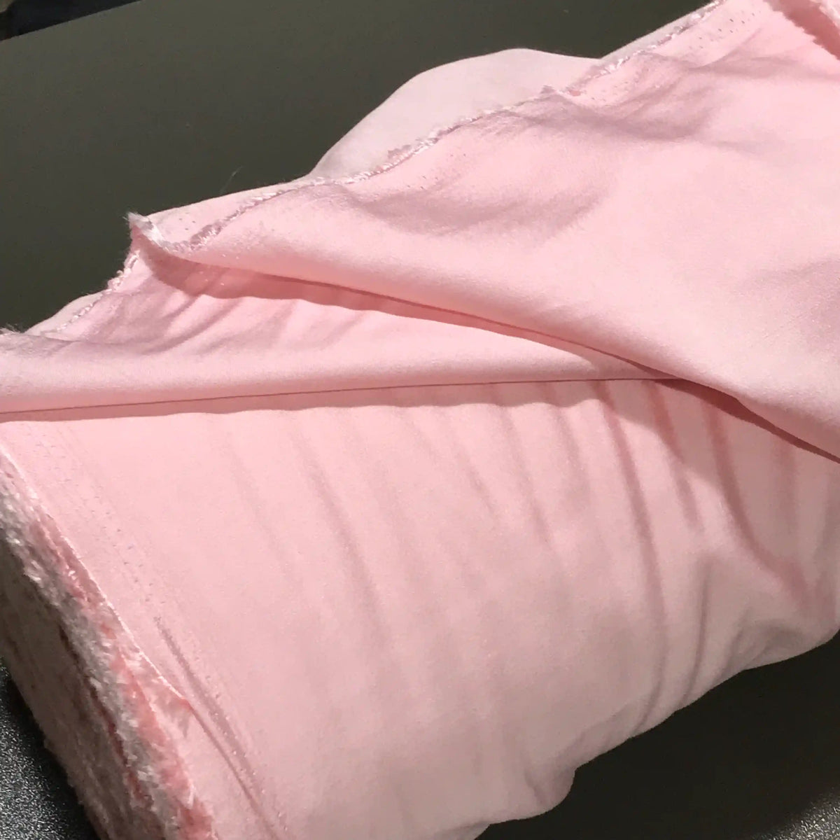 A close-up of a roll of Longan Craft’s Solid Color Ramie Fabric By The Yard in light pink, featuring rough, unfinished edges and partially unrolled on a dark surface. This super lightweight lyocell ramie material is breathable and showcases both texture and softness—ideal for blouses.