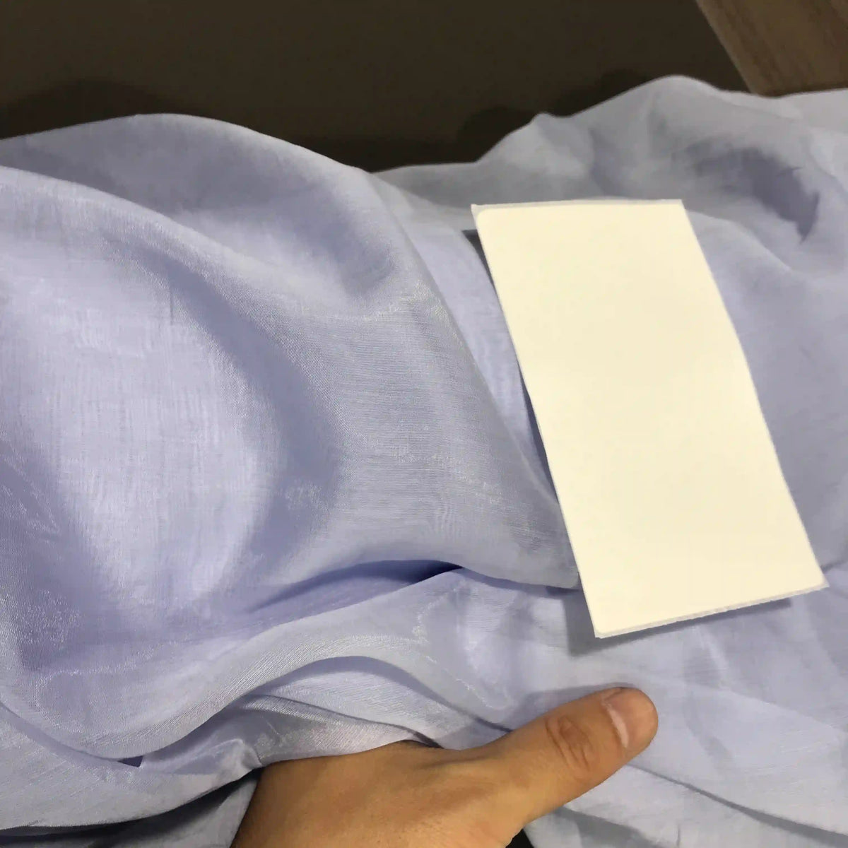 A hand holds the super lightweight light blue Solid Color Ramie Fabric by Longan Craft, showcasing its sheer, breathable texture with a rectangular white card placed on top to highlight the contrast in color and material.