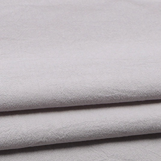 Close-up of folded Longan Craft Solid Color Washed Crepe Fabric by the Yard, made from 100% cotton. The light gray material features a soft, gently textured surface and a subtle crepe weave pattern, appearing smooth and neatly stacked—ideal for clothing or pants.