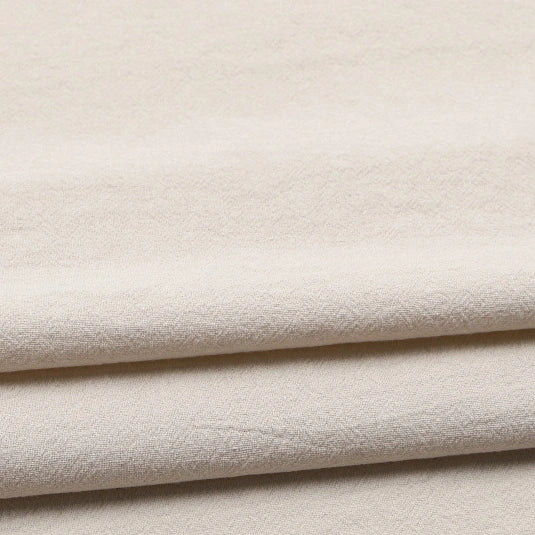 Three layers of light beige, softly textured Longan Craft Solid Color Washed Crepe Fabric are neatly stacked, forming gentle folds and delicate shadows. The surface displays a slightly rough texture, highlighting the 100% cotton crepe material’s natural feel and solid color finish—ideal for clothing and pants.