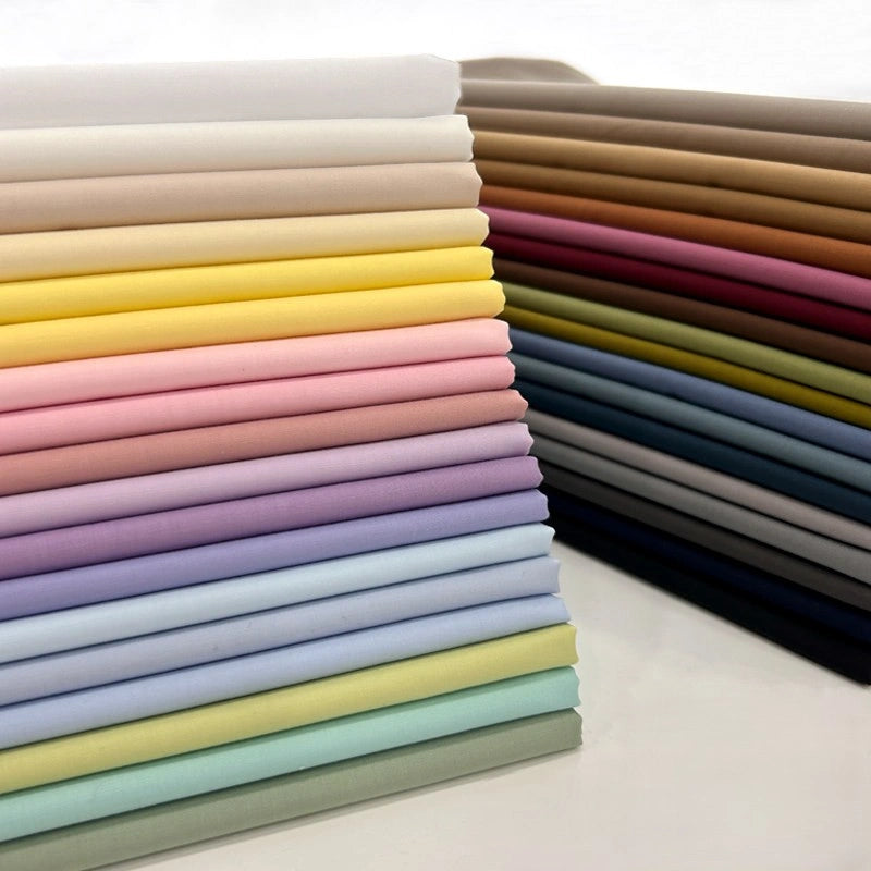 Neatly stacked Solid Cotton Lawn Fabric By The Yard from Longan Craft, in a range of colors—neutral to vibrant yellows, pinks, and blues. This polyester lawn cloth is ideal for crafting or tailoring and is displayed in rows on a white surface.