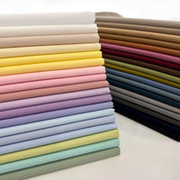 Neatly stacked Solid Cotton Lawn Fabric By The Yard from Longan Craft, in a range of colors—neutral to vibrant yellows, pinks, and blues. This polyester lawn cloth is ideal for crafting or tailoring and is displayed in rows on a white surface.