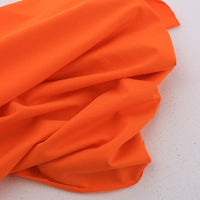 A piece of Longan Craft's Spandex Jersey Fabric By The Yard, in a solid bright orange, is displayed on a light surface with soft folds and wrinkles showcasing its four-way stretch and cotton jersey texture.