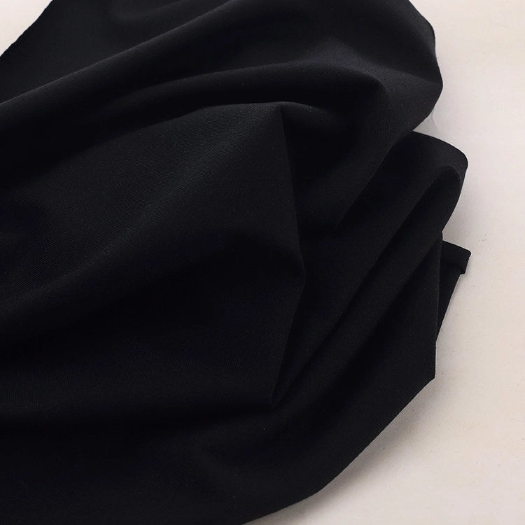 A close-up of Longan Craft's Spandex Jersey Fabric by the yard in black, draped in soft folds on a light surface. The solid color cotton jersey fabric features a smooth, matte finish and four-way stretch for comfort and flexibility.
