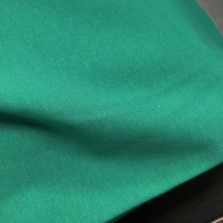 A close-up of Longan Craft's Spandex Jersey Fabric By The Yard in bright green, featuring subtle wrinkles, a soft texture, and four-way stretch as it rests on a dark surface.