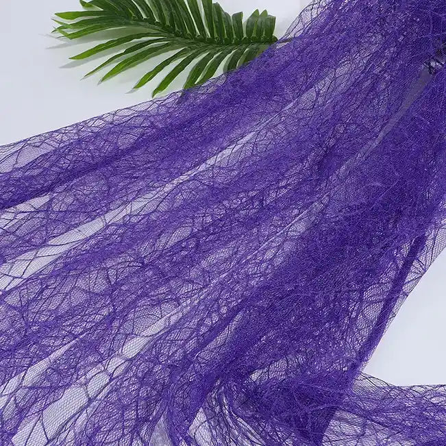 A piece of sheer, purple Spider Web Tulle Fabric By The Yard from Longan Craft is spread out on a white surface with a green palm leaf above—an ideal Halloween tulle fabric for DIY party dresses or festive decorations.