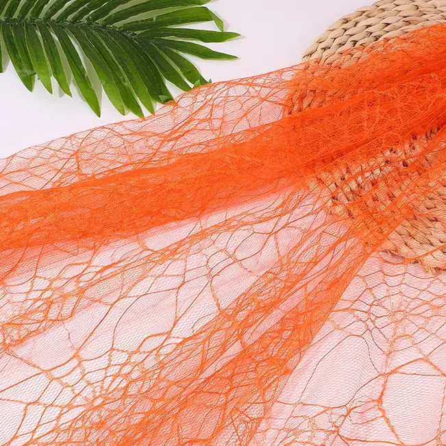 The Longan Craft Spider Web Tulle Fabric By The Yard in orange is displayed on a round woven mat with a green palm leaf—ideal for colorful Halloween DIY party dresses and festive décor.