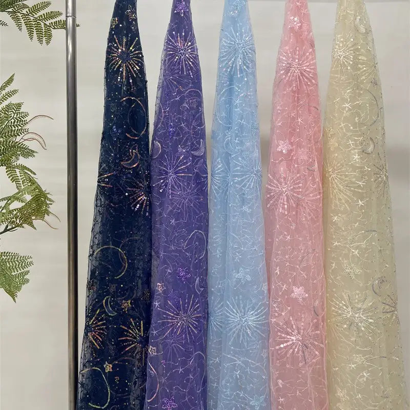 Five sheer Star and Moon Sequin Embroidered Tulle Fabrics by Longan Craft in navy, purple, light blue, pink, and ivory hang beside green ferns. Each features glittery stars, moons, and fireworks—perfect for DIY wedding dresses.
