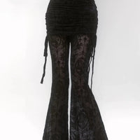 A person stands against a white background wearing a ruched black skirt with long ties over sheer bell-bottom pants made from Longan Craft’s Stretch Floral Spandex Tulle Fabric By The Yard, featuring ornate flocked patterns and crosses.