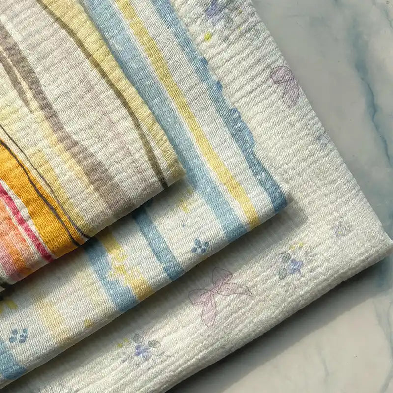 Three pieces of Longan Craft’s Striped Floral Print 100% Cotton Double Gauze Fabric are displayed on a marble surface. These breathable cloths feature pastel stripes and delicate floral or abstract prints in soft shades of yellow, blue, and purple, making them perfect for pajamas or other lightweight projects.