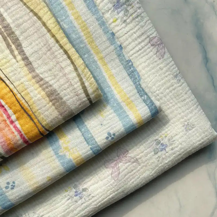 Three pieces of Longan Craft’s Striped Floral Print 100% Cotton Double Gauze Fabric are displayed on a marble surface. These breathable cloths feature pastel stripes and delicate floral or abstract prints in soft shades of yellow, blue, and purple, making them perfect for pajamas or other lightweight projects.