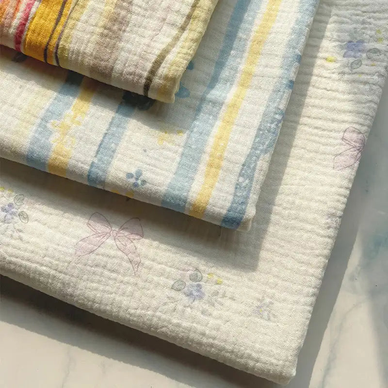 Four pieces of Longan Craft’s Striped Floral Print Gauze Fabric—crafted from 100% breathable cotton double gauze and adorned with pastel blue and yellow stripes, subtle bows, and delicate floral patterns—are neatly stacked on a light surface and gently illuminated by sunlight.