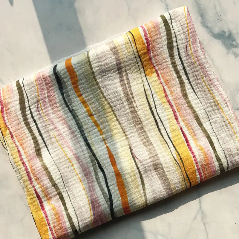 A folded piece of Longan Craft Striped Floral Print 100% Cotton Double Gauze Fabric, featuring a pattern of vertical stripes in yellow, orange, green, brown, pink, and gray tones, lies on a light marble surface with sunlight casting shadows—perfect material for pajamas.