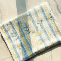 A piece of Longan Craft’s Striped Floral Print Gauze Fabric—100% cotton double gauze, featuring blue and yellow floral and stripe prints—lies folded on a light marble surface, softly illuminated by sunlight from one side.