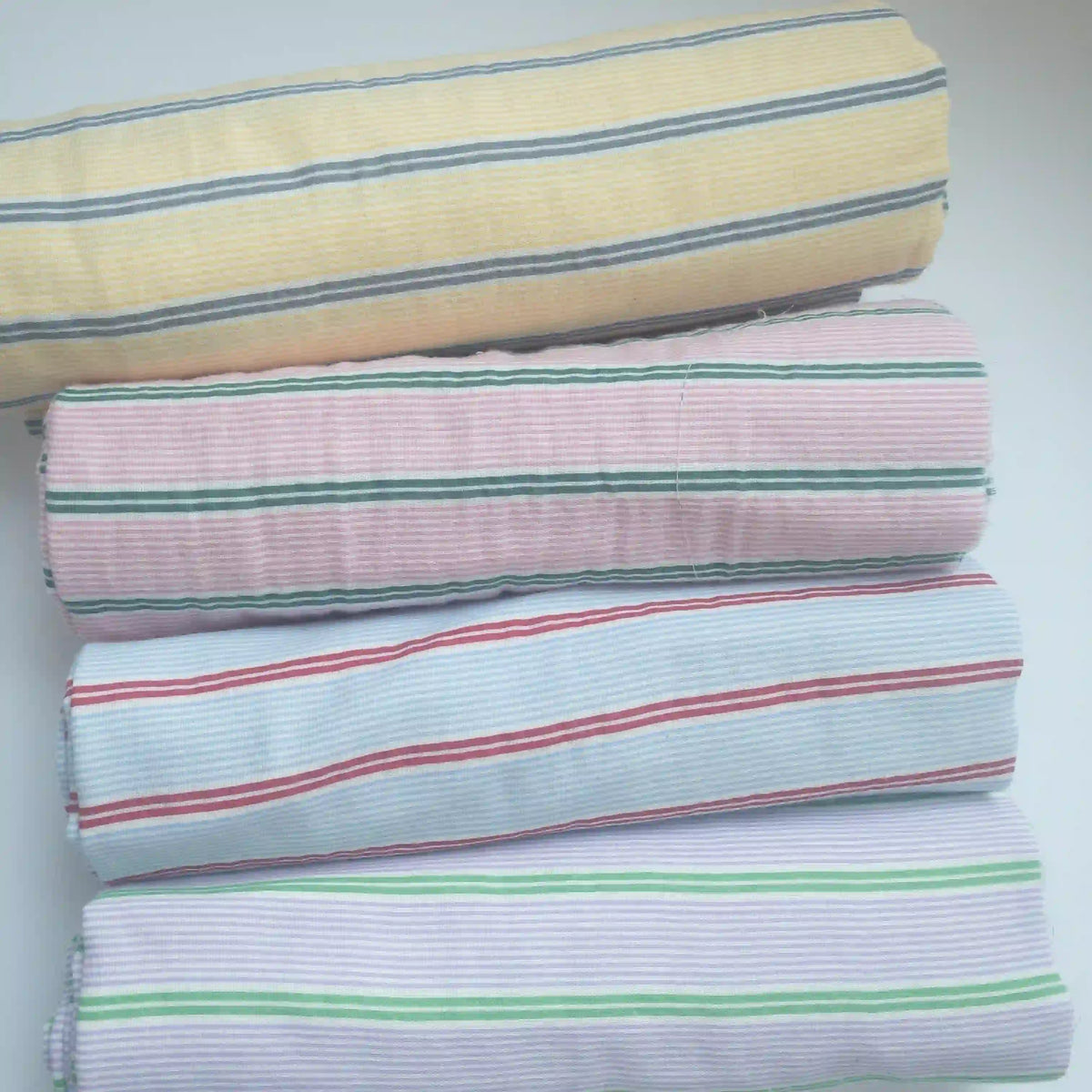 Four rolls of Longan Craft Striped Printed Gauze Fabric By The Yard—yellow/gray, pink/green, blue/red, lavender/green—are stacked horizontally. This double 100% cotton gauze is breathable and perfect for cozy, stylish projects.
